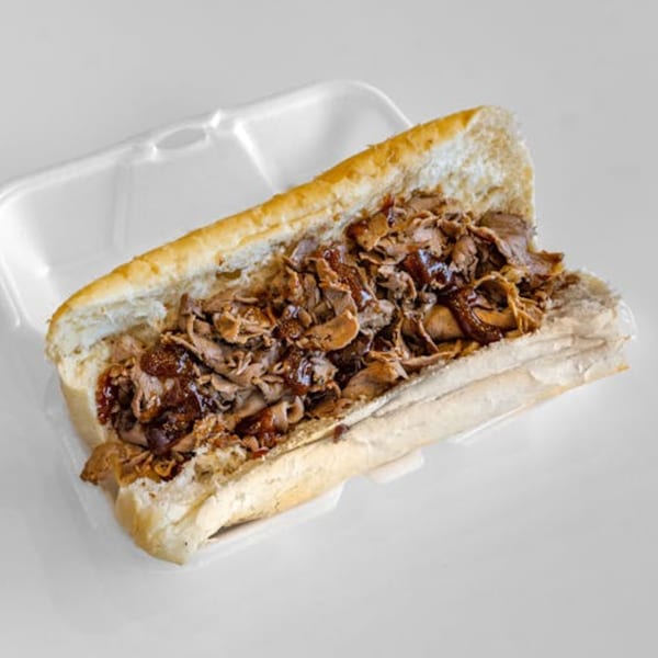 Unbeatable Eatables Inc. steak sandwich, with sliced meat and sauce on a long roll in a white container.