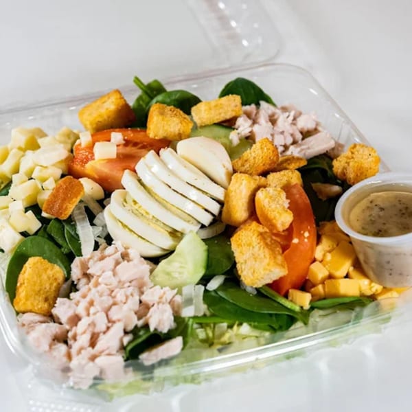 A colorful salad with chicken, cheese, tomatoes, eggs, and croutons in a clear plastic container.