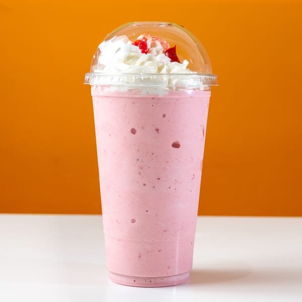 Strawberry milkshake with whipped cream and cherry topping in a clear plastic cup.