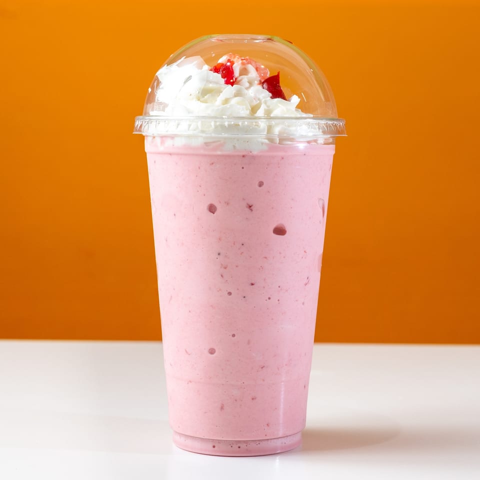 Strawberry milkshake with whipped cream and cherry topping in a clear plastic cup.