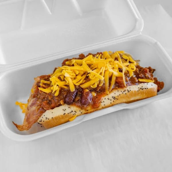 A chili cheese dog with poppy seeds on the bun, served in a white to-go container.