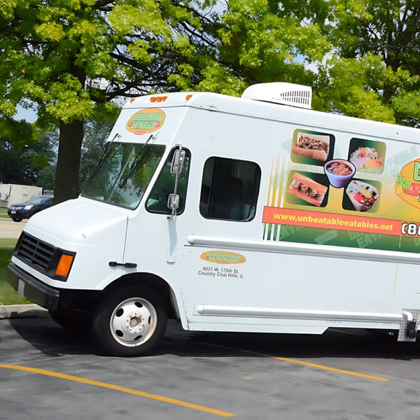 Unbeatable Eatables food truck, white with food images, parked on asphalt with green trees.